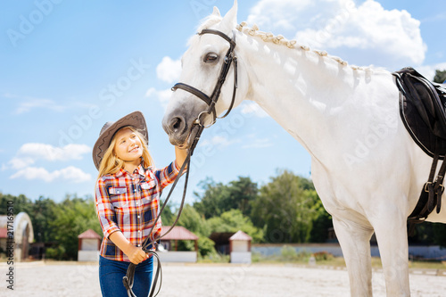 Beauty of horse. Cheerful blonde-haired girl wearing jeans and cowboy hat feeling amused by beauty of horse
