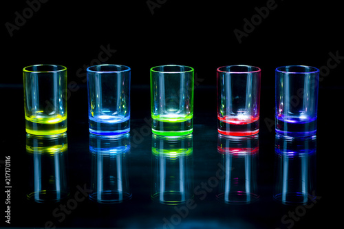 Five multicolored empty shot glasses reflecting on a glass surface symmetrically placed a black background © Алексей Торбеев