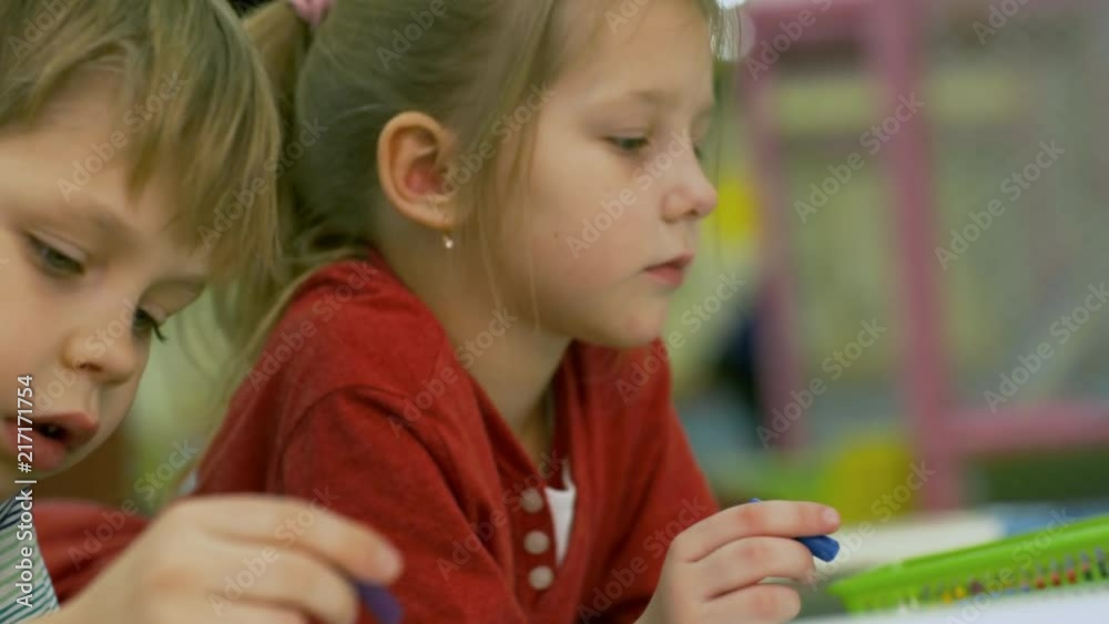 Close up faces of two cute children of primary school age drawing picture with colored wax chalks when lying on the floor Close up faces of two cute children of primary school age drawing picture with colored wax chalks when lying on the floor