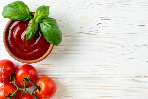 Tomato ketchup sauce in a bowl with basil and tomatoes on white wooden table. Top view. Copy space