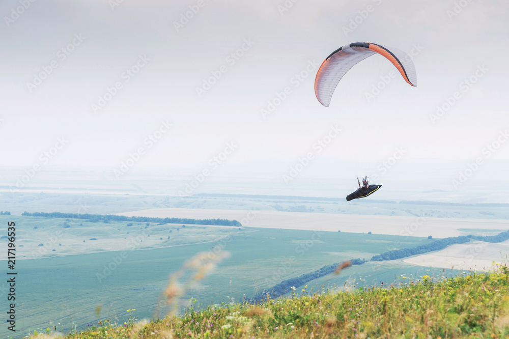 White orange paraglide with a paraglider in a cocoon against the background of fields of the sky and clouds. Paragliding Sports White orange paraglide with a paraglider in a cocoon against the background of fields of the sky and clouds. Paragliding Sports