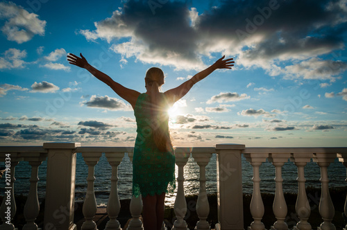 Silhouette of young caucasian woman with raised hands wearing green dress standing on balcony and looking at the sky and sea. Concept of fresh morning and happiness. photo