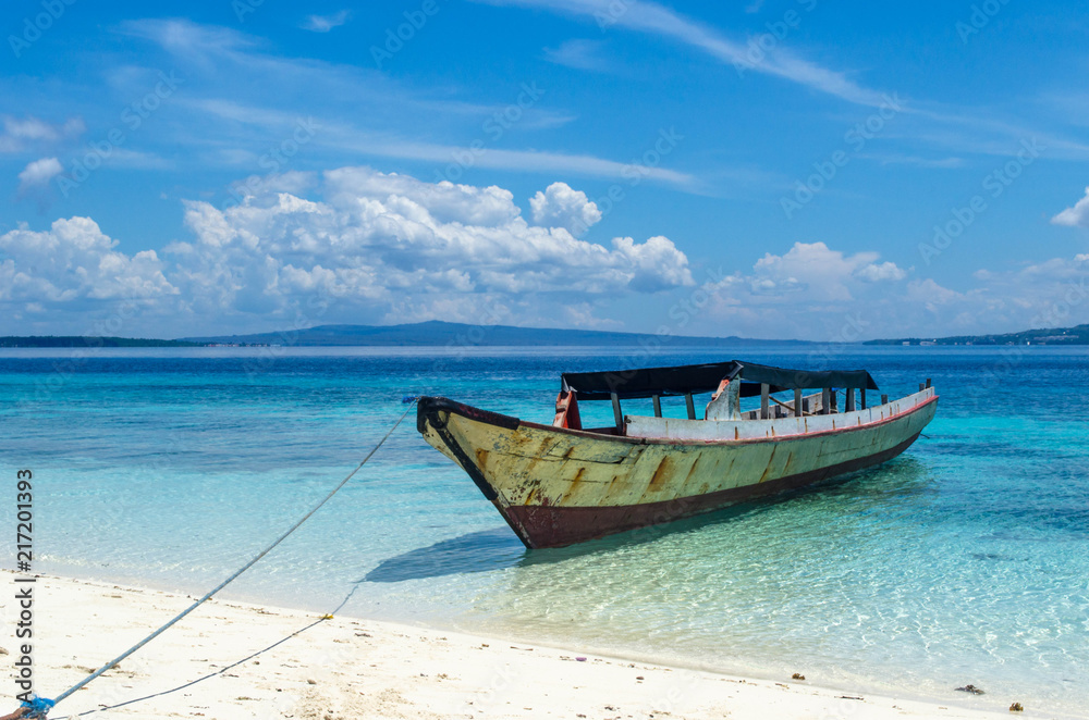 An isolated small boat is staying at the beach surrounded by tortoise clear water of the ocean. The sky is in white clouds. No people. Indonesia, Sulawesi An isolated small boat is staying at the beach surrounded by tortoise clear water of the ocean. The sky is in white clouds. No people. Indonesia, Sulawesi