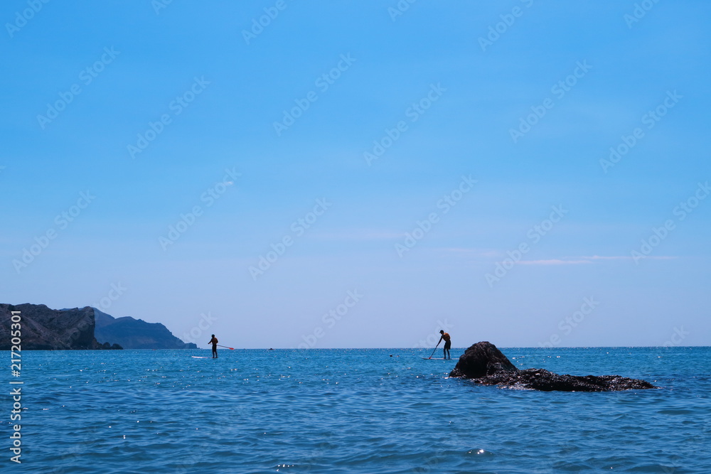 Fototapeta premium Walking along the sea while standing on a board with a paddle.