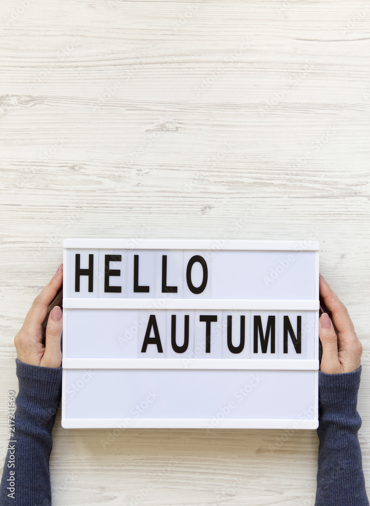 Female hands hold modern board with text 'Hello Autumn' over white wooden surface, top view. Overhead, flat lay, from above. Female hands hold modern board with text 'Hello Autumn' over white wooden surface, top view. Overhead, flat lay, from above.