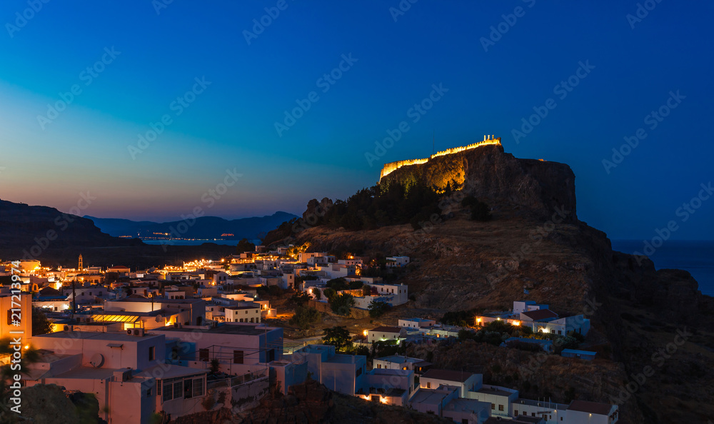 Scenic landscape photo of night Lindos town and castle on Rhodes island, Dodecanese, Greece. Panorama with bright lights, mountains and sea. Famous tourist destination in South Europe