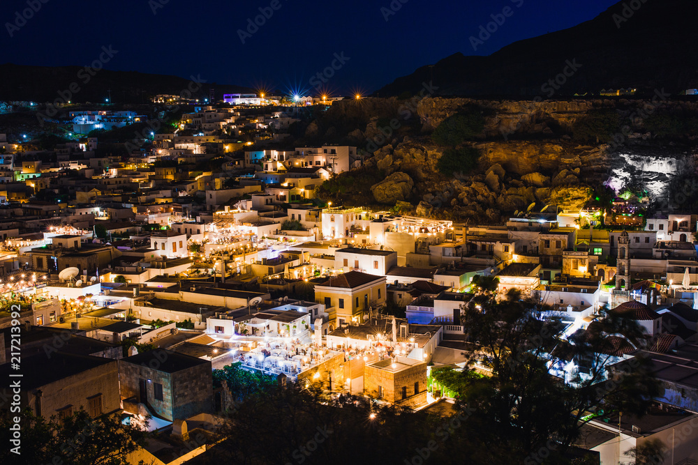 Obraz premium Scenic landscape photo of night Lindos town and castle on Rhodes island, Dodecanese, Greece. Panorama with bright lights, mountains and sea. Famous tourist destination in South Europe