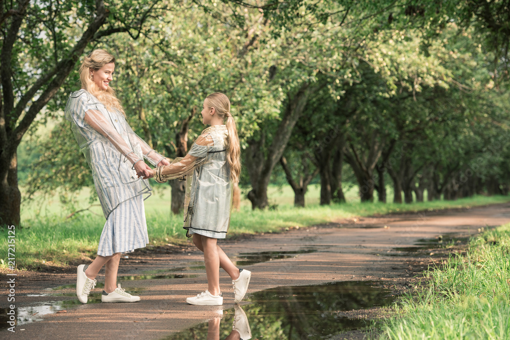 Fototapeta premium beautiful mother and adorable daughter in transparent raincoats holding hands on wet road in green park