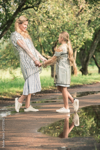 attractive mother and blonde daughter in transparent raincoats holding hands on wet road in park