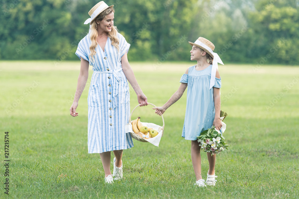 Fototapeta premium beautiful mother and daughter holding wicker basket with fruits and walking on green lawn