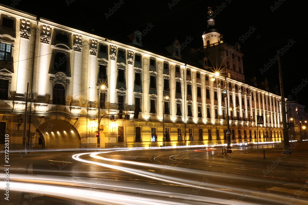Illuminated main building of the University of Wroclaw at night. A historic building in the Baroque style. Visible representative facade, tower and gate from the Odra river. Streaks of light from cars