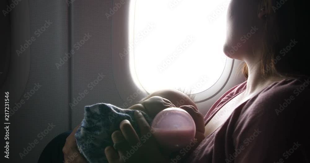 Mother looks out an airplane window holding a baby as she is drinking