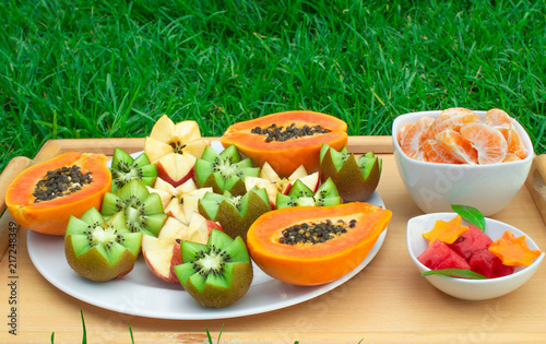 Still life on nature. Tray on the grass with papaya, kiwi, apples. Slices of mandarins and watermelon in plates.