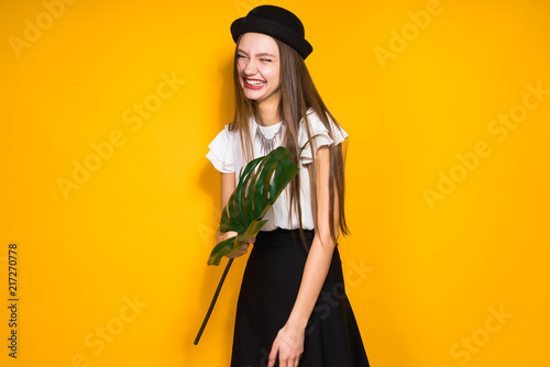 laughing stylish girl model in black hat posing against yellow wall background, holding a green leaf in hands