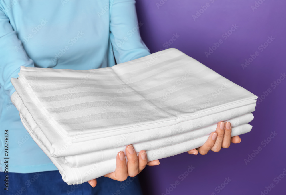 Woman holding stack of bed linen on color background, closeup