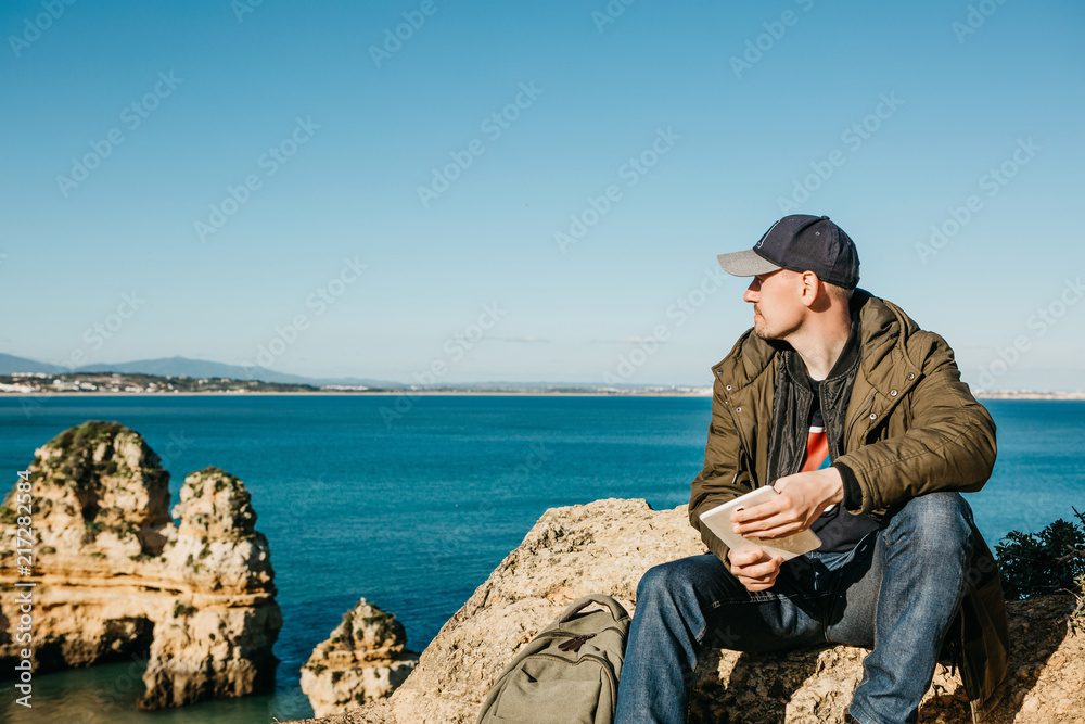 A young man or boy or tourist uses a tablet and looks afar into the beautiful Atlantic Ocean off the coast of Portugal next to the city called Lagos.