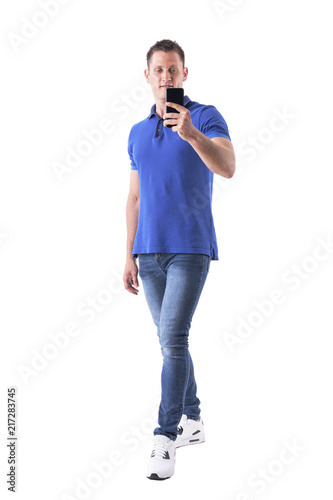 Happy young adult casual man in blue polo shirt taking selfie with smart phone. Full body isolated on white background. 