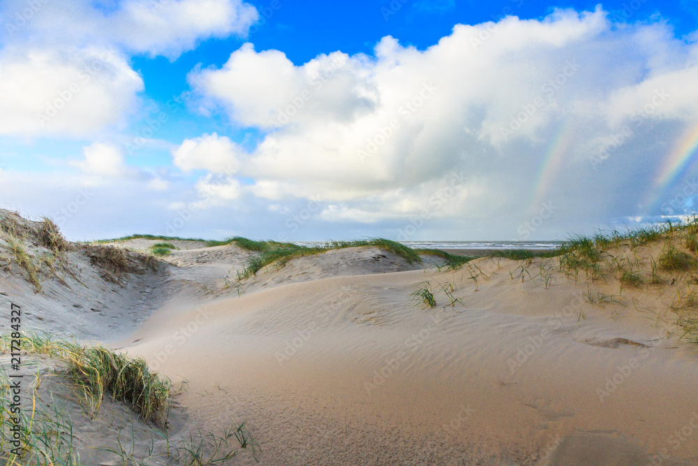 Newly formed Dunes on Dutch North Sea coast at IJmuiderslag with wind sweeping stripes in the sand against the background of blue sky with scattered clouds and double rainbow Newly formed Dunes on Dutch North Sea coast at IJmuiderslag with wind sweeping stripes in the sand against the background of blue sky with scattered clouds and double rainbow