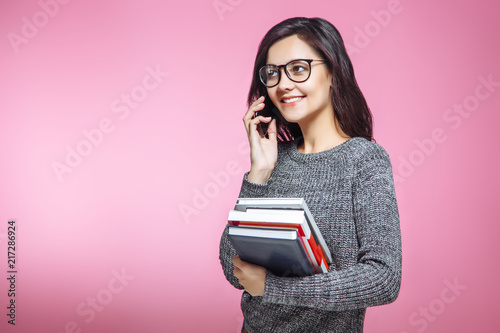 Cute female student holding books when talking on the phone on pink background