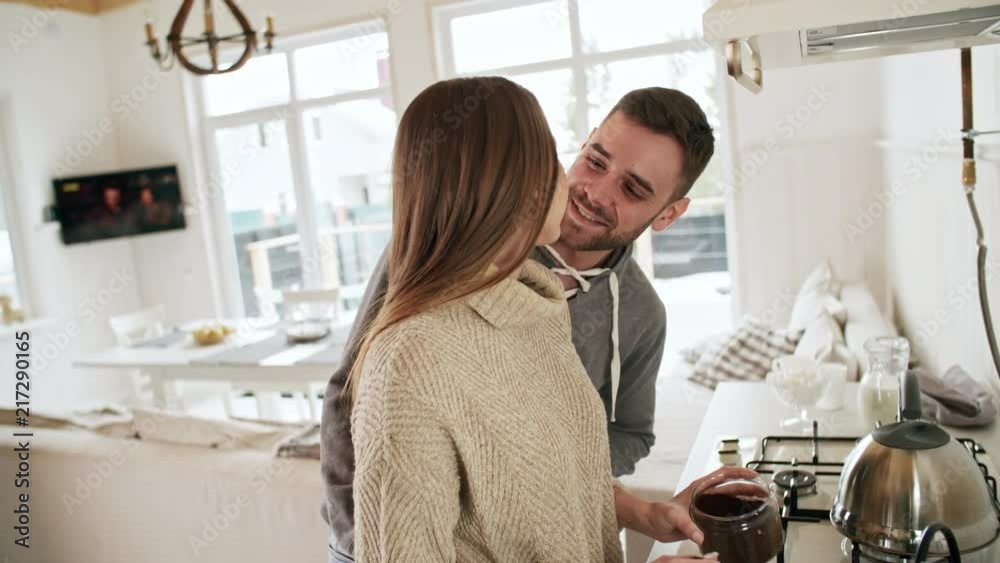 Handheld shot of romantic man with beard walking up to his beautiful young girlfriend making coffee in kitchen: he is kissing her on cheek and talking Handheld shot of romantic man with beard walking up to his beautiful young girlfriend making coffee in kitchen: he is kissing her on cheek and talking