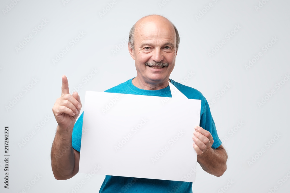 Mature bald man in blue tshirt holding blank sheet of paper, smiling Mature bald man in blue tshirt holding blank sheet of paper, smiling