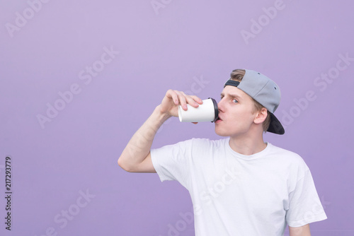 Funny student in a white T-shirt and cap drink coffee from a glass on a purple background and looking at one eye in the camera.Portrait a young man drinking coffee at the background of a purple wall