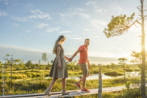 side view of couple holding hands while walking on wooden bridge with green plants and blue sky on background