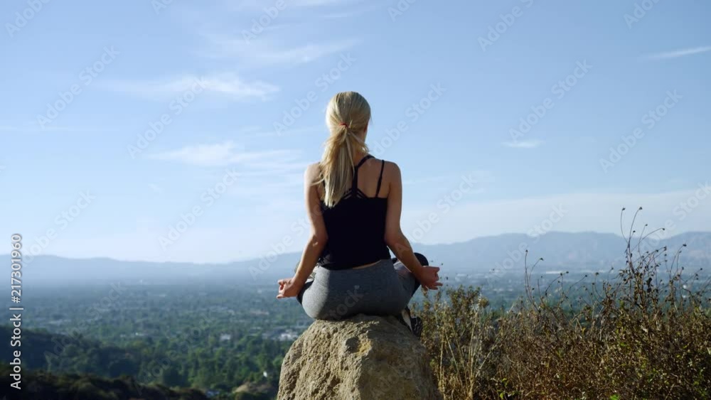 Yong blonde woman seen from behind sitting on a rock outside in yoga pose with blue sky and beautiful backdrop views Yong blonde woman seen from behind sitting on a rock outside in yoga pose with blue sky and beautiful backdrop views