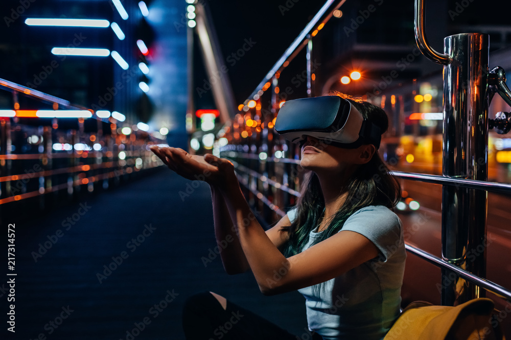 young woman in virtual reality headset sitting on street with night city on background