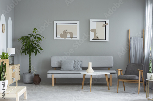 Posters above grey sofa in minimal living room interior with plants and armchair. Real photo