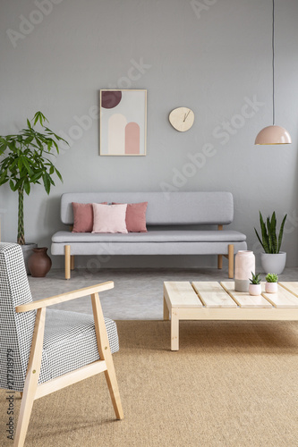 Armchair and wooden table in grey living room interior with poster and clock above sofa. Real photo