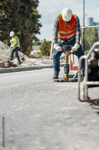 Worker in helmet and reflective vest with drill repairing asphalt