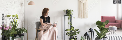 Young woman covered with blanket sitting on armchair and reading a book in white apartment interior with many fresh plants