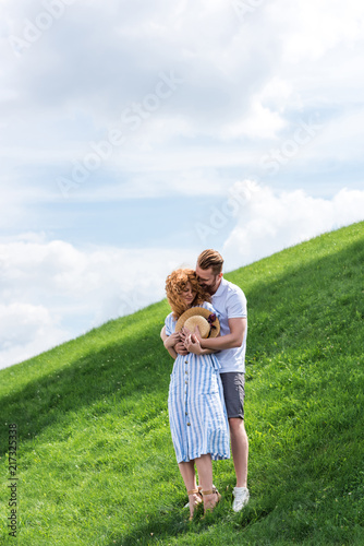 smiling redhead man hugging girlfriend from behind on green hill