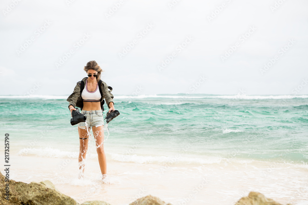 Hipster fit woman in sexy shorts, khaki shirt, sunglasses and travel backpack pour out water from army boots. Modern tourist traveler, active lifestyle, sport adventure camp and extreme on vacation. Hipster fit woman in sexy shorts, khaki shirt, sunglasses and travel backpack pour out water from army boots. Modern tourist traveler, active lifestyle, sport adventure camp and extreme on vacation.