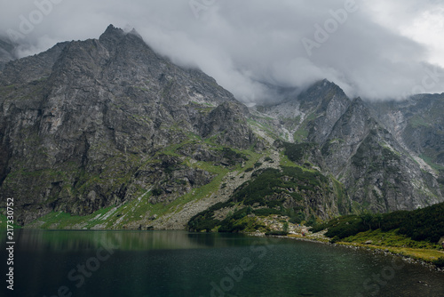 Beautiful view of foggy mountains cover by dark clouds and green forest with a reflection in a lake. Morskie Oko. Marine Eye. High Tatras  Zakopane  Poland