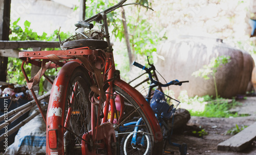 Old red steel vintage bicycle is parked in very old garage with many other unused stuff such as bamboo, kid bike, used bottle and water tank