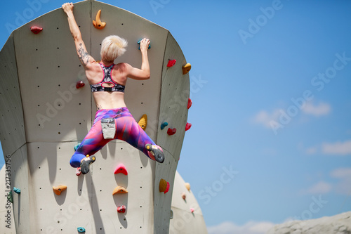 Photo from back of young athlete blonde with short hairstyle climbing wall for rock climbing against blue sky