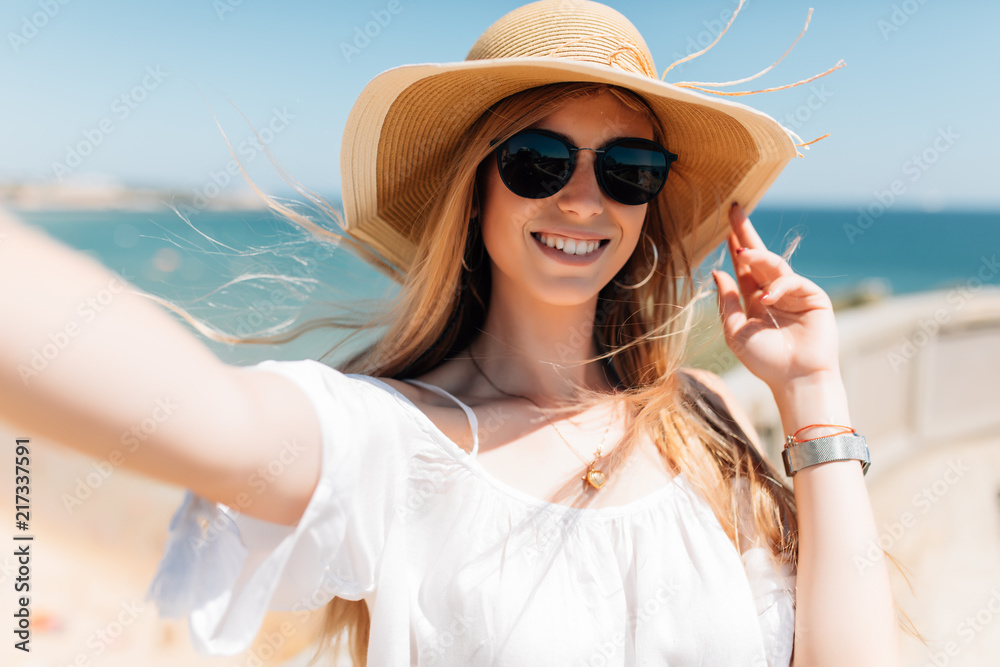 Fototapeta premium Portrait of happy adorable woman making selfie or photo of herself, stands against sea background. People and leisure concept