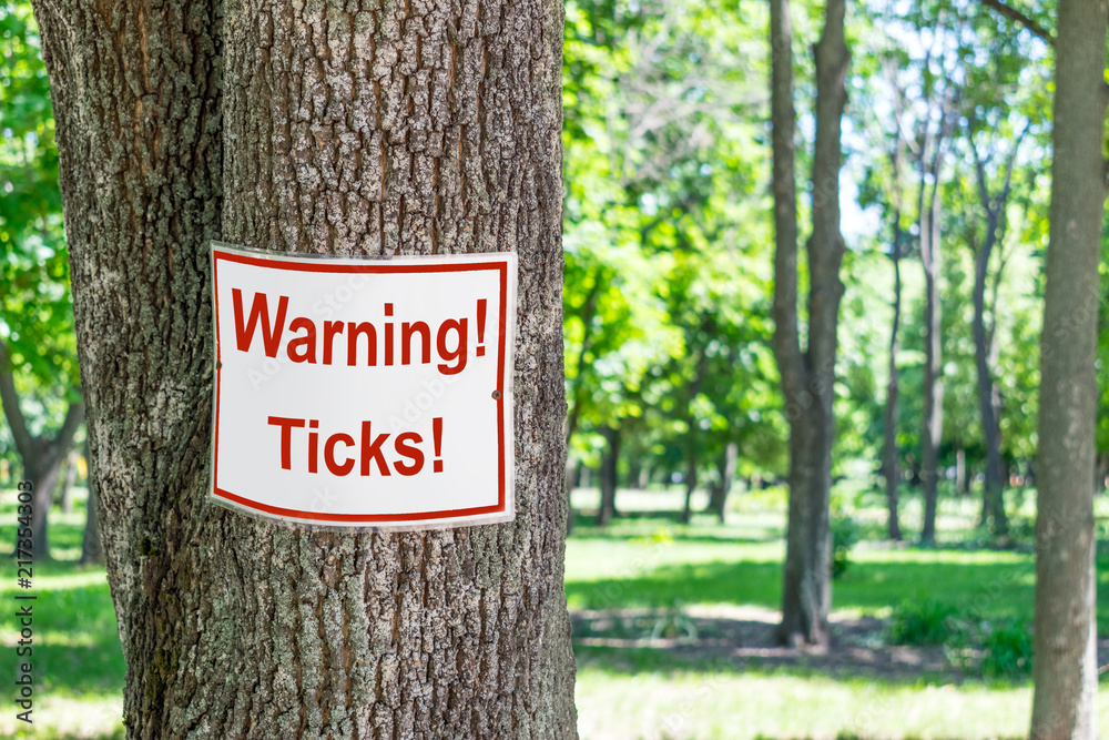 Sign Warning ticks on tree in green park background. White and red