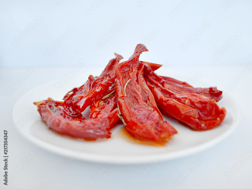 Red homemade oven dried tomatoes with rosemary on a white saucer against a light background, close-up, front view Red homemade oven dried tomatoes with rosemary on a white saucer against a light background, close-up, front view