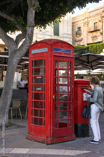 Woman send postcard via red post box near english red phone booth in Valletta