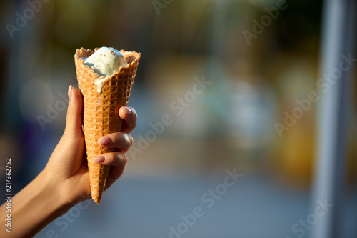 Ice cream cone in woman hand with blurred city bokeh background and copyspace. Isolated close view, selective focus. Icecream melting and defrosting. Summer theme. © artiemedvedev