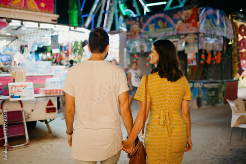 Pretty brunette girl with her boyfriend with beard walking holding hands with their backs at the fair in Spain in the evening