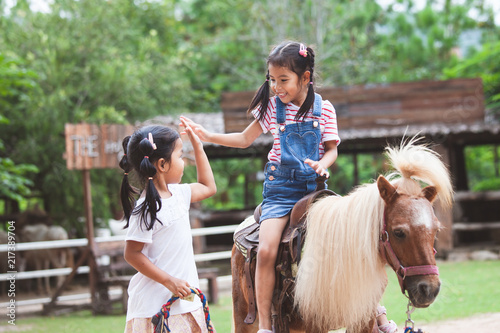 Canvas Print Cute asian child girl riding a pony and making hi five gesture with her elder si