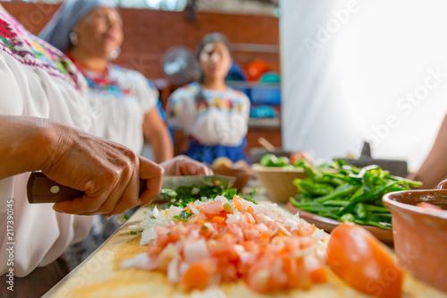 Mujeres en preparación de alimentos, picando jitomate, cebolla y chiles sobre una tabla en una cocina tradicional mexicana