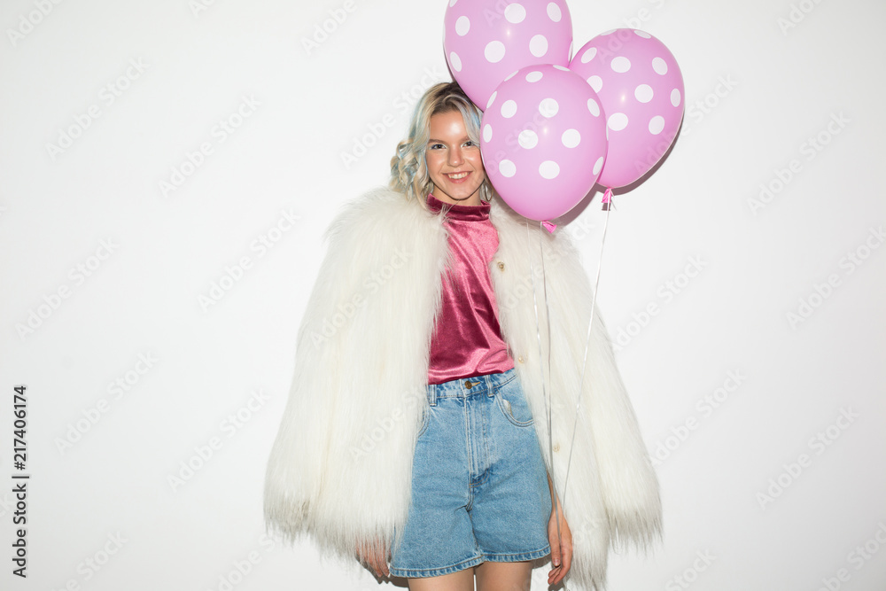 Pretty smiling girl in white fur jacket and velvet blouse happily looking in camera while holding pink balloons in hand over white background Pretty smiling girl in white fur jacket and velvet blouse happily looking in camera while holding pink balloons in hand over white background