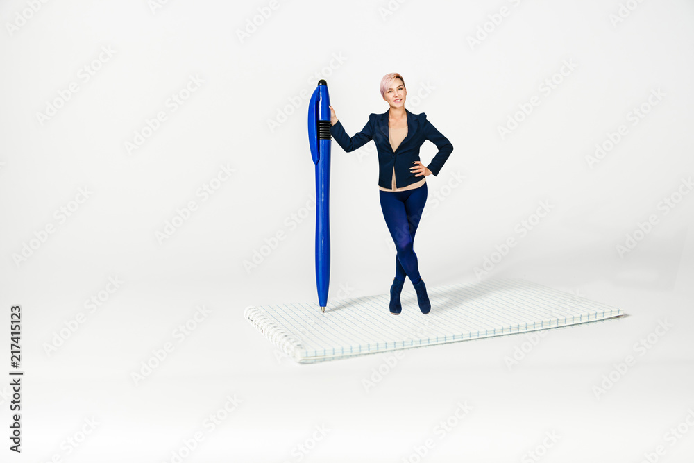 Young beautiful girl office worker stands holding on to huge pen and notebook on a white background . Young beautiful girl office worker stands holding on to huge pen and notebook on a white background .