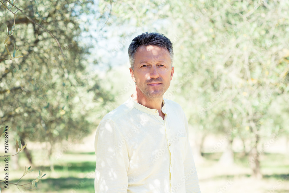 Handsome man posing in olive trees garden. Male portrain over mediterranean olive field ready for harvest. Confident mature man in italian olive's grove with ripe fresh olives. Handsome man posing in olive trees garden. Male portrain over mediterranean olive field ready for harvest. Confident mature man in italian olive's grove with ripe fresh olives.