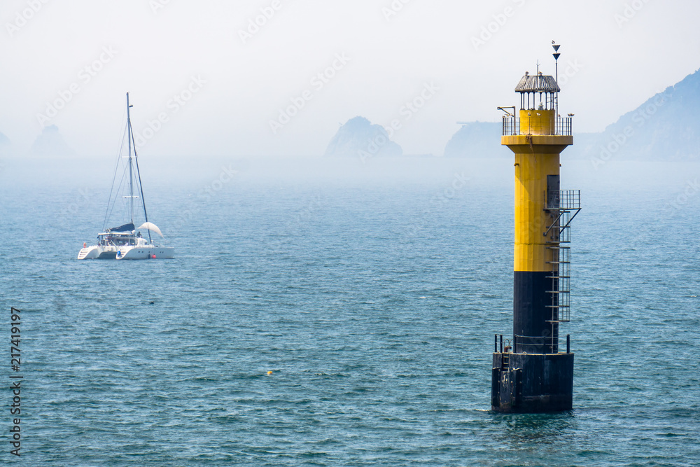 Yellow black lighthouse standing in the sea. There are several seagulls at the top. And small catamaran sail boat anchor near by.